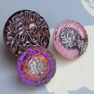 May include: Three decorative glass buttons in shades of purple, pink, and iridescent colors. The buttons have unique textures and designs, including a swirling pattern and a floral embellishment. These buttons are ideal for sewing and craft projects.