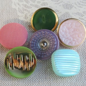 May include: A selection of six decorative buttons in diverse colours and designs. The buttons include a textured pink button, a green button with gold accents, a purple button with a silver centre, a light pink button with a floral design, a light blue button with horizontal lines, and a green button with gold text.