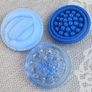 May include: Three blue buttons with different designs. One is light blue with a ribbed pattern, another is a darker blue with a beaded design, and the third is a clear blue with a faceted pattern. These buttons are suitable for sewing and craft projects.