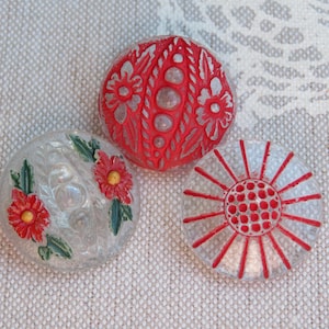 May include: Three decorative buttons with floral designs. One is red with a carved floral pattern. Another is clear with red and yellow flowers. The third is clear with a red sunburst design.