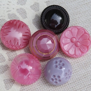 May include: A collection of six round buttons in various shades of pink, purple, and black. The buttons feature distinct textures including geometric facets, floral petal patterns, radial lines, and small circular indentations on their surfaces.