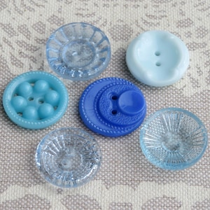 May include: A collection of six vintage buttons in shades of blue. The buttons vary in design, including smooth, textured, and patterned surfaces. The colors range from light blue to a deep cobalt, offering a variety of options for sewing and crafting projects.