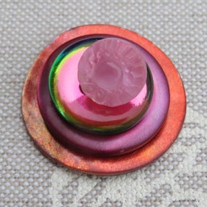 May include: A layered button with a stack of circular elements. The base is a reddish-orange, followed by a deep purple, a shimmering iridescent layer, and a small, textured pink button on top. The button is a decorative craft item.