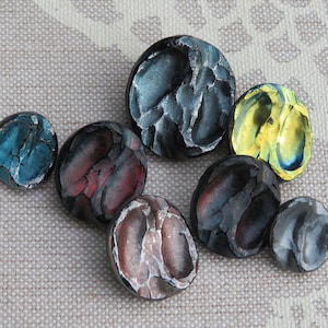 May include: A selection of artisan buttons in various sizes and colours. The buttons have a textured, stone-like appearance with hues of blue, grey, red, and yellow. The buttons are arranged on a neutral, textured surface.