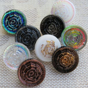 Könnte beinhalten: Eine Sammlung runder, dekorativer Knöpfe in verschiedenen Farben, darunter schillernd, schwarz, weiß und bronze. Jeder Knopf hat ein Spiraldesign. Geeignet für Näh- oder Bastelprojekte.