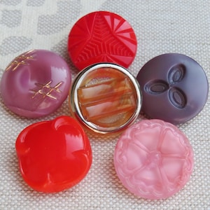 May include: A collection of six round buttons in various colors and designs. The set includes a bright red geometric button, a mauve button with gold hash marks, a central amber button with a silver rim, a dark purple button, a red button, and a pink floral button.