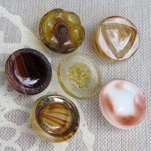 Op de afbeelding: Een verzameling van zes ronde knopen met diverse ontwerpen. Eén is een donker amberkleurige bloemvorm, een andere is doorschijnend beige met een driehoekspatroon, een donkerbruine gestreepte knoop, een textuurrijke gele glazen knoop en een wit met roze werveling.