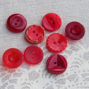 May include: Eight red buttons of different shapes and designs. Some have two holes, others a shank. The buttons are made of a glossy material and are arranged on a textured, light-coloured surface. Measurements are not available.