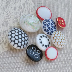 May include: A collection of nine white, black, blue, red, and gold buttons with various patterns and designs. The buttons are arranged on a beige fabric background.