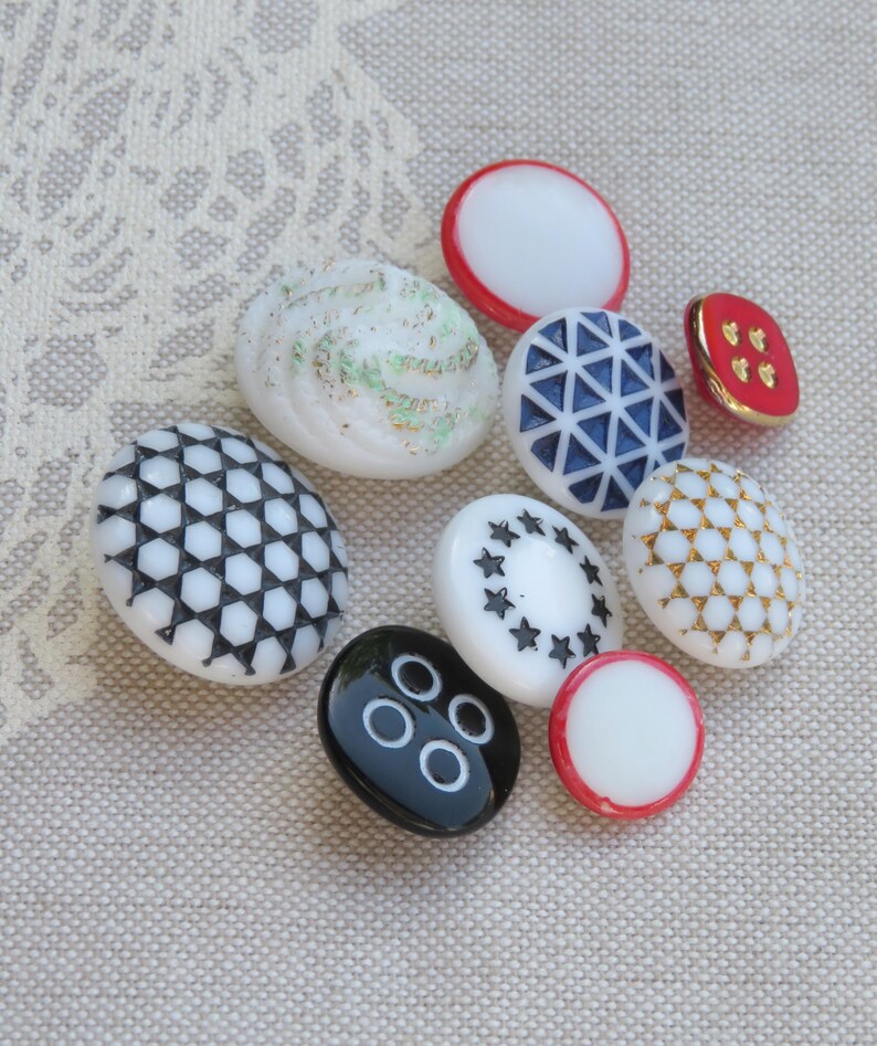 May include: A collection of nine white, black, blue, red, and gold buttons with various patterns and designs. The buttons are arranged on a beige fabric background.