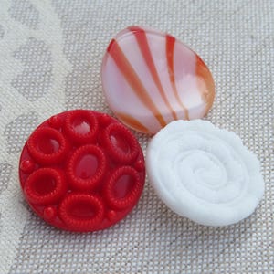 May include: Three circular buttons: one bright red button with a textured pattern of raised oval shapes, one white button with a swirling spiral relief design, and one translucent button featuring alternating white and orange-red stripes.