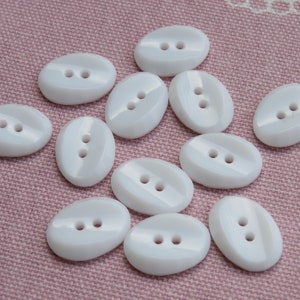 Puede incluir: Una colección de botones ovalados blancos con dos agujeros cada uno. Los botones están dispuestos en un patrón aleatorio sobre un fondo de tela rosa.