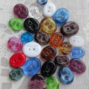 Op de afbeelding: Een verzameling kleurrijke, ovaalvormige knopen in verschillende kleuren, waaronder rood, blauw, groen en bruin. De knopen hebben twee gaten en een decoratief ontwerp. Sommige zijn transparant, andere ondoorzichtig.