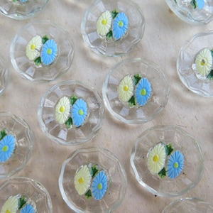 Op de afbeelding: Transparante glazen decoratieve knopen met een geschulpte rand. Elke knoop heeft een geschilderd ontwerp van twee madeliefjes, één geel en één blauw, met groene bladeren. De knopen zijn gerangschikt op een lichtgekleurd oppervlak.