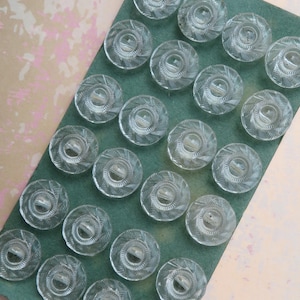 May include: A collection of clear, circular buttons with a textured, spiral design. The buttons are arranged on a green rectangular surface, with a light pink and beige background. These buttons are ideal for sewing and craft projects.