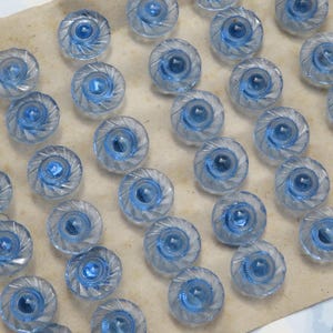 May include: A collection of clear, spiral-designed buttons with a central blue accent. The buttons are arranged on a light brown surface, showcasing their intricate details and reflective qualities. These buttons are ideal for sewing and craft projects.