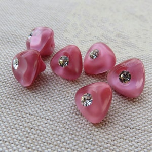 May include: Six pink heart-shaped buttons with a clear rhinestone in the centre. The buttons are on a white fabric background.