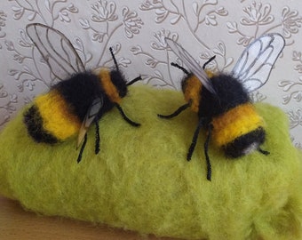 May include: Two needle-felted bumblebee figures with black and yellow fuzzy bodies, thin black wire legs, and translucent white wings with black vein details, resting on a textured, bright lime green felted wool block.
