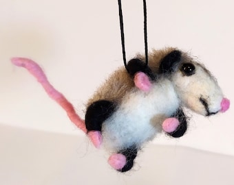 May include: A needle-felted opossum ornament featuring a white and tan fuzzy body, black limbs, a long pink tail, and a pink nose. The small animal is suspended by a thin black string attached to its back, creating a hanging display piece.