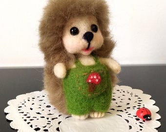 May include: A needle-felted hedgehog figurine with brown fur, wearing green overalls featuring a small red mushroom appliqué on the front. The hedgehog has black eyes, a black nose, and a pink mouth. A small red ladybug figurine sits nearby.