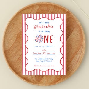 Puede incluir: Una tarjeta de invitación festiva con un borde a rayas rojas y blancas y un borde festoneado. El texto dice "our little firecracker is turning ONE" con detalles del evento. La tarjeta está colocada sobre un plato de madera.