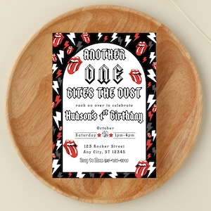 May include: A birthday invitation with a black, white, and red design. The invitation features the text "ANOTHER ONE BITES THE DUST" and "Hudson's 1st Birthday." The design includes lightning bolts and the Rolling Stones' tongue logo.