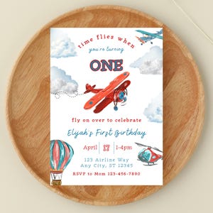 May include: A watercolor-style invitation for a first birthday celebration. The design features a red airplane, a hot air balloon, and a helicopter, with clouds and the text "time flies when you're turning ONE". The invitation includes the date, time, and location.