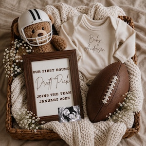 May include: A brown teddy bear wearing a white football helmet, a cream-colored long-sleeve baby bodysuit with the text Baby Andersen, a brown wooden frame with the text Our First Round Draft Pick Joins The Team January 202X, and a brown football.