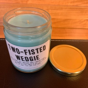 K&ouml;nnte beinhalten: Eine tealblaue Kerze in einem Glasgef&auml;&szlig; mit goldenem Deckel. Das Etikett auf dem Gef&auml;&szlig; lautet "Two-Fisted Wedgie".