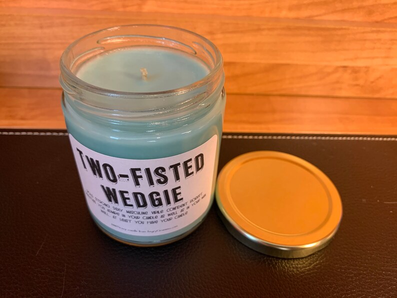 K&ouml;nnte beinhalten: Eine tealblaue Kerze in einem Glasgef&auml;&szlig; mit goldenem Deckel. Das Etikett auf dem Gef&auml;&szlig; lautet "Two-Fisted Wedgie".