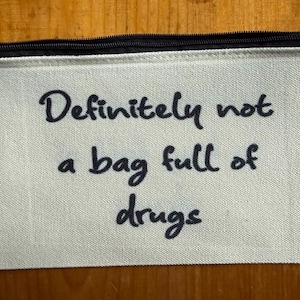 May include: A rectangular off-white canvas zipper pouch featuring the black printed text "Definitely not a bag full of drugs" in a casual, handwritten-style font. The pouch has a black zipper closure along the top edge and a textured fabric.
