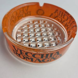 May include: Orange glass ashtray with a black inscription that reads "Vecchia Romagna".