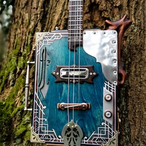 Puede incluir: Una guitarra de caja de puros azul turquesa con una placa de protección plateada y detalles dorados. La guitarra tiene un mástil de madera y un diapasón decorativo. La guitarra está apoyada en un tronco de árbol.