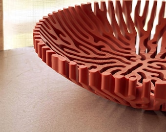 Può includere: Una ciotola color terracotta caratterizzata da una complessa struttura a reticolo ramificato simile al corallo. Il design è composto da numerose nervature verticali interconnesse che si irradiano dal centro, creando vuoti di forma organica.