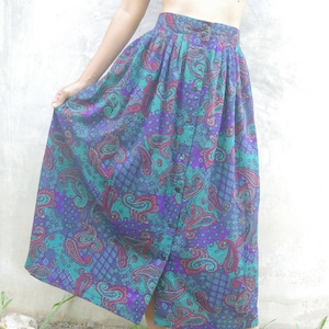 Op de afbeelding: Een lange, zwierige rok met een paisley print in tinten turquoise, paars en bordeauxrood. De rok heeft een knoopsluiting aan de voorkant en een geplooide taille.