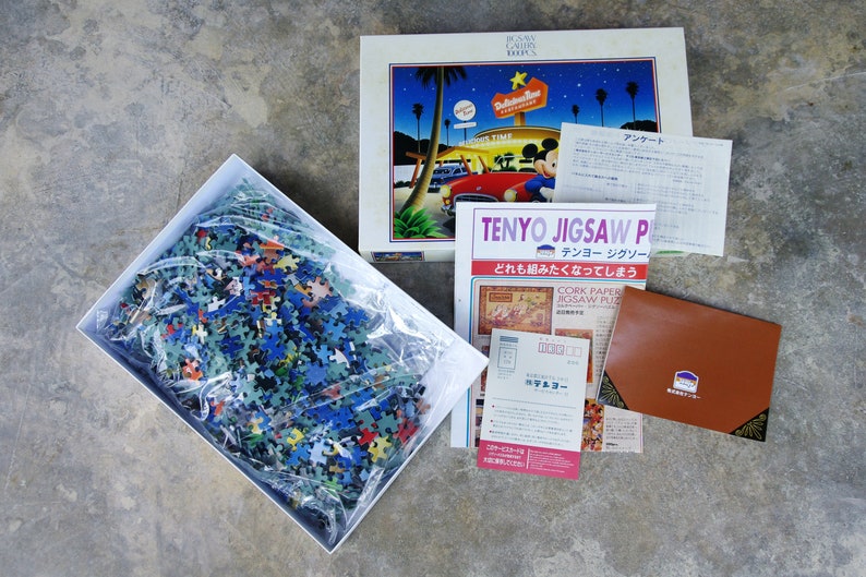 May include: A complete 1000-piece jigsaw puzzle in a white box with a clear plastic bag containing the puzzle pieces. The puzzle is of a cartoon scene with Mickey Mouse and a diner. The box has the text "TENYO JIGSAW PUZZLE" and "CORK PAPER JIGSAW PUZZLE" on it.
