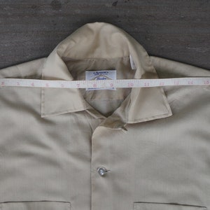 May include: A beige button-up shirt with a collar and a pocket on the left side. The shirt has a label that reads "Armoire" and "Studio". A measuring tape is placed across the top of the shirt, showing a measurement of 16 inches.