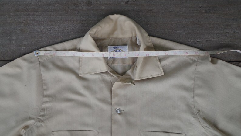 May include: A beige button-up shirt with a collar and a pocket on the left side. The shirt has a label that reads "Armoire" and "Studio". A measuring tape is placed across the top of the shirt, showing a measurement of 16 inches.