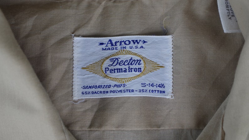 May include: A white tag with blue text that reads "Arrow> Made in U.S.A. Decton Permafron Sanforized-Plus S-14-14 1/2 65% Dacron Polyester-35% Cotton"