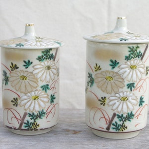 Puede incluir: Dos tazas de té de cerámica blanca con tapas, que presentan un diseño floral de crisantemos blancos y beige con hojas verdes sobre un fondo crema.