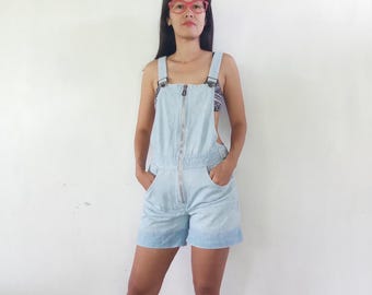 Puede incluir: Pantalones cortos de peto vaqueros azul claro con cremallera y bolsillos. Las correas se aseguran con cierres metálicos. La modelo lleva un top estampado debajo del peto. Los pantalones cortos son una prenda informal.