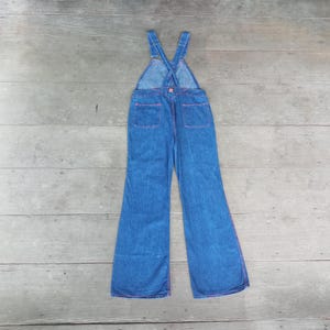 Könnte beinhalten: Blaue Jeans-Latzhose mit ausgestelltem Bein-Design. Die Latzhose hat rosa Nähte um die Taschen und Nähte. Die Träger kreuzen sich am Rücken und sind mit einem rosa Knopf befestigt.