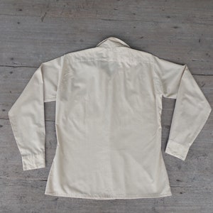 May include: A long-sleeved, light cream colored button-down shirt with a collar.