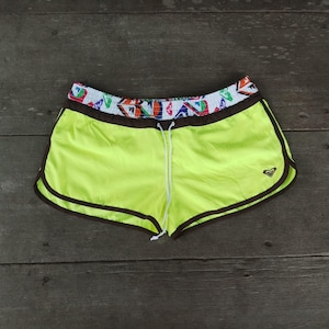 May include: Neon yellow athletic shorts with a drawstring waist and brown trim. The waistband features a colorful geometric pattern. A small logo is visible on the lower right leg. The shorts are made of a lightweight material.