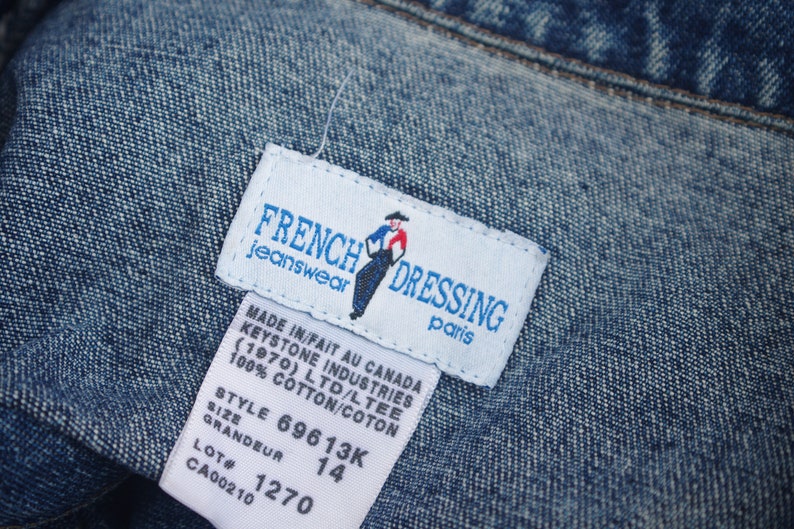 May include: White tag with blue text and a small illustration of a person wearing a cowboy hat. The tag reads "French Dressing Jeanswear Paris" and includes the text "Made in/Fait au Canada Keystone Industries (1970) Ltd/Ltee 100% Cotton/Coton Style 69613K Grandeur 14 Lot # 1270 CA00210".