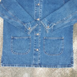 May include: A blue denim jacket with a button-up front and two large pockets on the front. The jacket has long sleeves with buttoned cuffs.