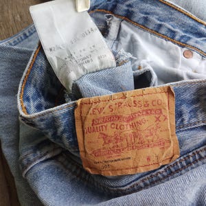 Puede incluir: Primer plano de unos vaqueros de mezclilla azul claro con un parche de cuero marr&oacute;n que dice "Levi Strauss & Co. Original Riveted Quality Clothing". Una etiqueta blanca con texto est&aacute; adjunta a los vaqueros. Los vaqueros est&aacute;n doblados.