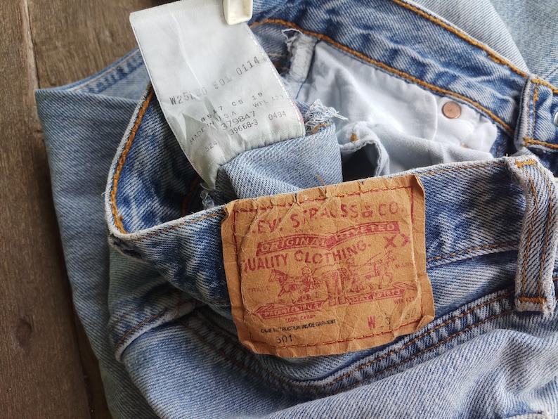 Puede incluir: Primer plano de unos vaqueros de mezclilla azul claro con un parche de cuero marr&oacute;n que dice "Levi Strauss & Co. Original Riveted Quality Clothing". Una etiqueta blanca con texto est&aacute; adjunta a los vaqueros. Los vaqueros est&aacute;n doblados.