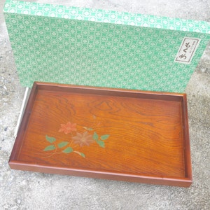 Puede incluir: Una bandeja de servir de madera con un diseño floral en tonos verdes, naranjas y marrones. La bandeja está en una caja con un patrón verde y blanco y la palabra japonesa "さくら" impresa en el lateral.