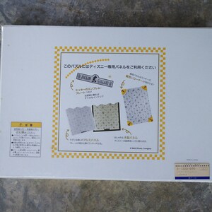 May include: White box with a yellow and black checkered border. The box features a picture of a jigsaw puzzle with a Mickey Mouse design. The text on the box reads "Jigsaw Gallery" and "This puzzle uses Disney-exclusive panels".